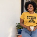 Woman in a mustard Running on Spite and Coffee tee holding a coffee cup outside a cafe